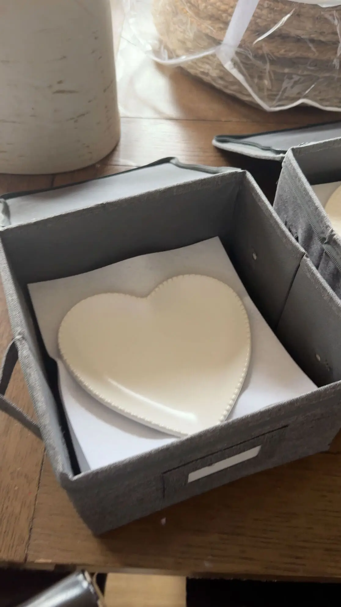 A white ceramic heart-shaped plate with a beaded edge carefully stored in a gray fabric storage box with padded dividers, sitting on a wooden table.